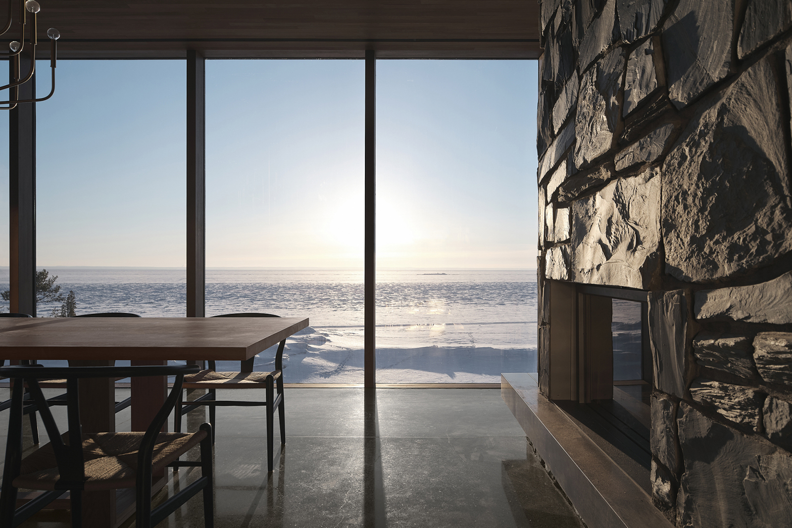 Salle de repos vitrée possédant un foyer de pierre et une table de bois donnant sur une vallée de neige
